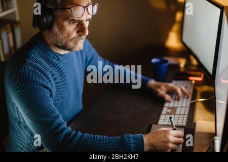 Uomo maturo con cuffie sedute alla scrivania a casa che lavora con tablet grafico e computer Foto Stock