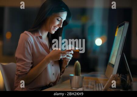 Giovane donna sorridente seduto alla scrivania in ufficio utilizzando uno smartphone Foto Stock
