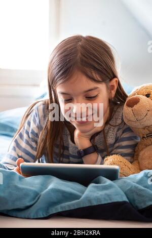 Ritratto di una ragazza sorridente che si stette a letto con orsacchiotto utilizzando un tablet digitale Foto Stock