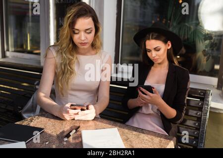 Le amiche adorabili usano gadget seduti sulla veranda estiva al bar Foto Stock