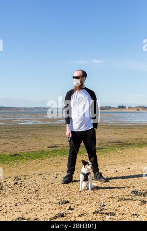 Uomo che indossa una maschera protettiva del viso cammina il suo cane lungo una spiaggia nelle sue ore di permesso quotidiano per l'esercizio che il governo ha permesso durante il co Foto Stock