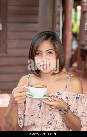 Mano donna asiatica holding Hot caffè Espresso sormontato da uno a forma di cuore il latte in vetro bianco. Foto Stock