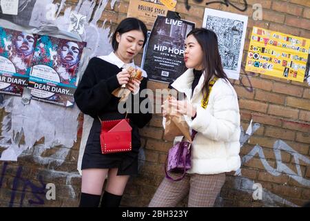 Due ragazze asiatiche di fronte a un muro di mattoni che mangia cibo, Brick Lane, Londra Foto Stock
