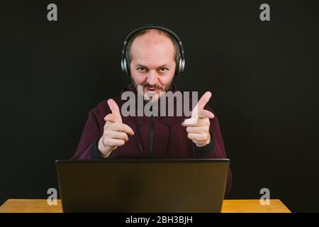 Ragazzo con felpa e cappuccio seduto dietro la scrivania con computer portatile e cuffie su sfondo nero, parlare con il microfono, registrare video blog Foto Stock