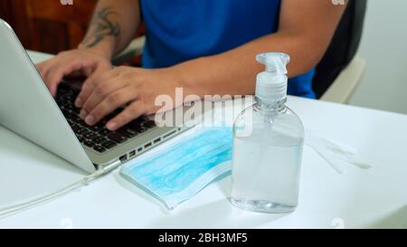 Avvicinati a una bottiglia di plastica di gel di alcol accanto a una maschera blu sul viso di un tavolo bianco accanto a un uomo in una camicia blu seduto a lavorare con il suo laptop Foto Stock
