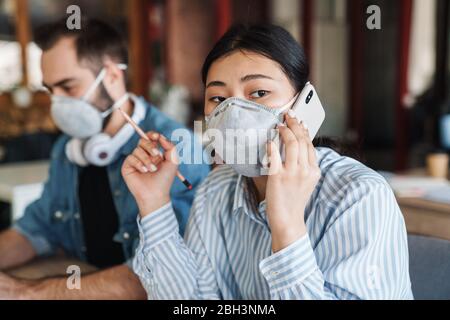 Foto di giovani studenti multinazionali in maschere mediche che parlano al cellulare mentre studiano in classe Foto Stock