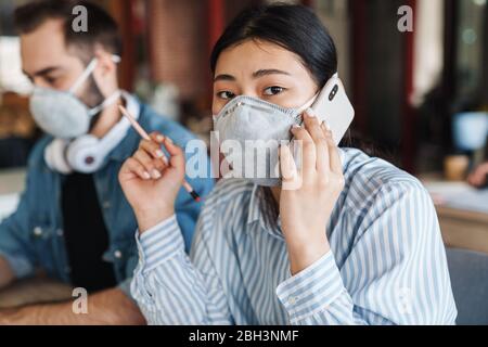 Foto di giovani studenti multinazionali in maschere mediche che parlano al cellulare mentre studiano in classe Foto Stock