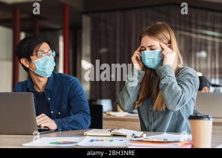Foto di giovani studenti multinazionali con mal di testa in maschere mediche che studiano su laptop in classe Foto Stock