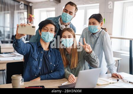 Foto di studenti allegri multinazionali in maschere mediche prendendo selfie sul cellulare in classe Foto Stock