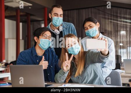 Foto di studenti allegri multinazionali in maschere mediche prendendo selfie sul cellulare in classe Foto Stock