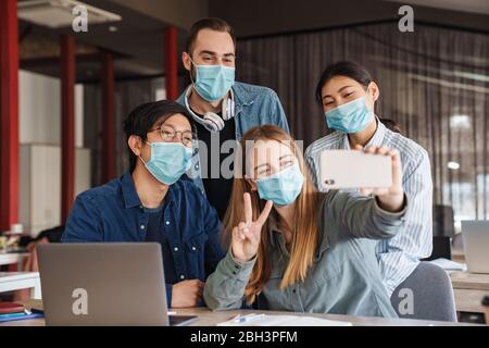 Foto di studenti allegri multinazionali in maschere mediche prendendo selfie sul cellulare in classe Foto Stock