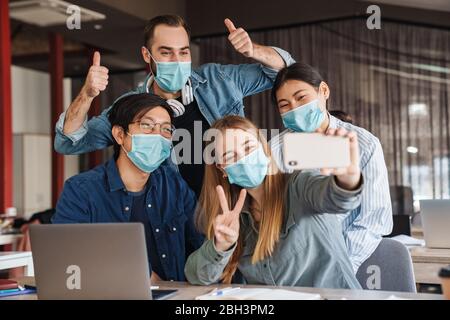 Foto di studenti allegri multinazionali in maschere mediche prendendo selfie sul cellulare in classe Foto Stock