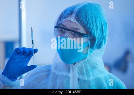 Medico femminile in costume protettivo che tiene la siringa con il medicinale in mano mentre lavora in ospedale Foto Stock