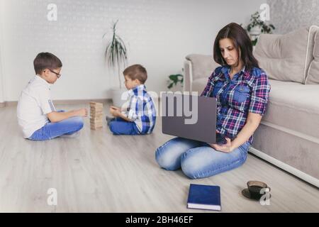 Rimani a casa, mamma che lavora in remoto su un notebook nel soggiorno mentre i bambini giocano. Compiti a casa. Concetto di distanza sociale. Quarantena. Chiudi, Foto Stock