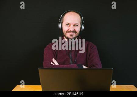 Ragazzo con felpa e cappuccio seduto dietro la scrivania con computer portatile e cuffie su sfondo nero, parlare con il microfono, registrare video blog Foto Stock