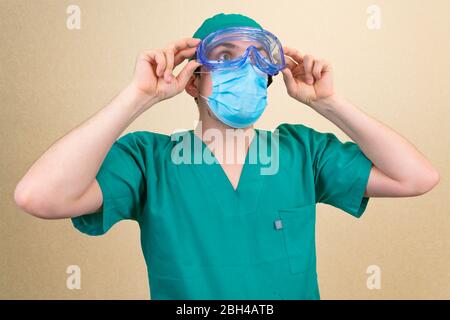 Medico in tuta verde e maschera blu e occhiali protettivi su sfondo giallo. Foto Stock