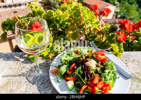 Primo piano di bicchiere di vino bianco e piatto di insalata in giardino con fiori di geranio rosso all'esterno in giardino d'estate in Toscana Foto Stock