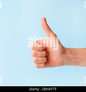 Primo piano della mano femminile che mostra i pollici su segno contro sfondo blu pastello, spazio copia, concetto minimo Foto Stock