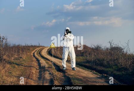 Istantanea orizzontale dell'uomo in tuta bianca, maschera a gas e guanti in piedi sulla strada del villaggio in campo accanto al segno tossico che mostra il dito medio, zona di radiazione. Concetto di ecologia, pericolo e pericolo. Foto Stock