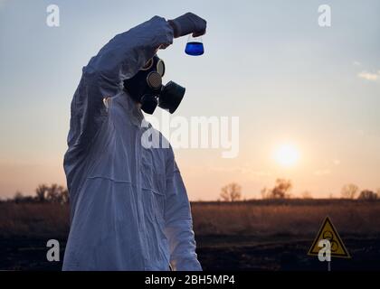 Scienziato in tuta bianca, maschera a gas e guanti che tengono il tubo di prova con liquido blu mentre studiano campioni di terreno su territorio bruciato con segno di rischio biologico, facendo lavori di ricerca sul campo con erba bruciata Foto Stock