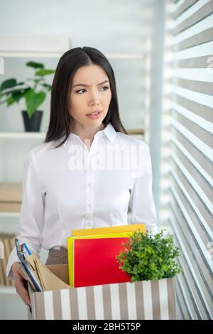 Al lavoro. Sconvolto giovane femmina brunetta guardando nella finestra, tenendo la scatola con il suo personale roba Foto Stock