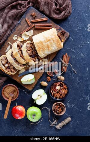 Strudel di mele di pasta filo servito su un tagliere con ingredienti su fondo di cemento scuro: Zucchero marrone, noci pecan, bastoncini di cannella, uva passa Foto Stock