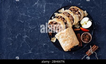 Rotolo di strudel di mele caramellate avvolto in pasta fillo con noci, cannella, uvetta servita su un piatto nero con anice stellato, su un piatto scuro Foto Stock