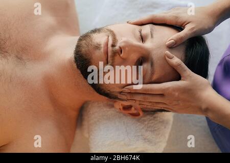 Uomo affascinante che si rilassa con gli occhi chiusi durante il massaggio alla testa presso il centro benessere. trattamento antidolorifico per la testa con massaggio Foto Stock