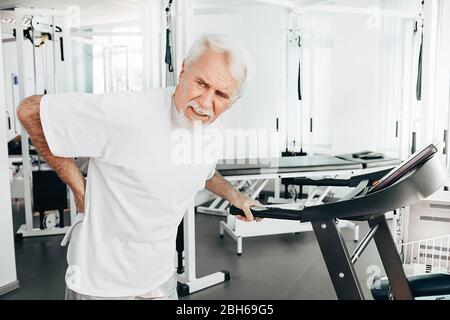 Uomo anziano che soffre di mal di schiena. Stare in piedi vicino alla pedana mobile e sentirsi male Foto Stock