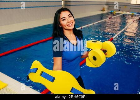 Donna sportiva che tiene in mano i barcampanelli fitness in piscina e guarda positivamente la macchina fotografica. Istruttore di Aqua fitness Foto Stock