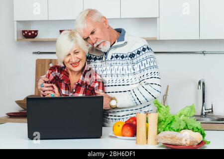 Anziani, buona coppia, cucina insieme, utilizzando il laptop mentre si sta in piedi in una cucina bianca, a casa. Cucinare sano mangiare per le persone anziane Foto Stock
