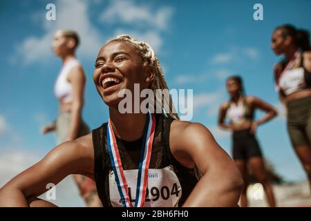 Atleta femminile sorridente dopo aver vinto una gara con altri concorrenti in background. Sportivo con medaglia che celebra la sua vittoria allo stadio. Foto Stock