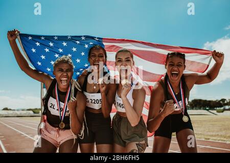 Gruppo di runner femminili con medaglie vincenti una competizione. Le atlete americane celebrano la vittoria mentre si levano insieme sulla pista che tiene il Foto Stock