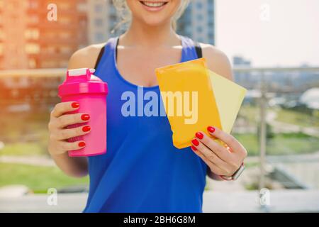 donna sportiva che tiene in mano uno shaker e bustine con un cocktail proteico solubile. nutrizione sportiva per allenamenti intensivi all'aperto. Foto Stock