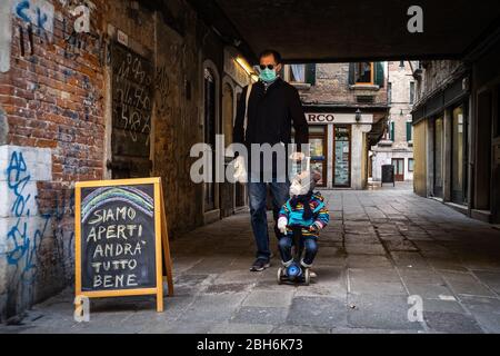 VENEZIA, ITALIA - APRILE 2020: Un uomo e suo figlio, indossando maschere protettive, camminano accanto ad un cartello con scritto 'siamo aperti, tutto andrà bene' d Foto Stock