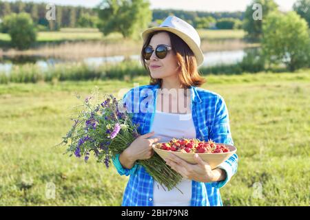 Contadina con ciotola di fragole appena raccolte e bouquet di fiori selvatici. Sfondo della natura, paesaggio rurale, prato verde, stile country Foto Stock