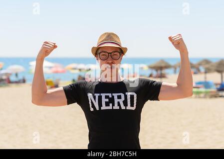 Giovane uomo turistico nerd flettendo entrambe le braccia alla spiaggia Foto Stock