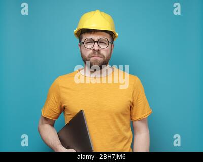 Uomo o ingegnere sospettoso in un casco da costruzione e occhiali rotondi con un laptop e guarda con gli occhi schiacciati. Foto Stock