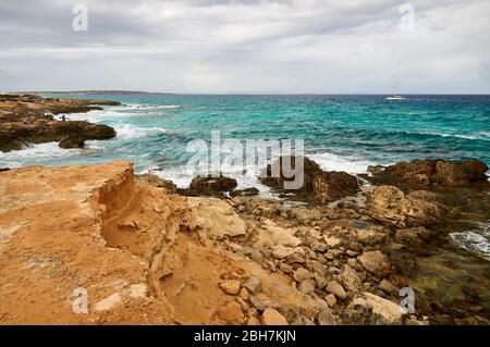 Pescatore sulle rocce e barca a vela lungo la costa di es Carnatge con acque azzurre e ruvide vicino ad es Caló (Formentera, Isole Baleari, Mar Mediterraneo, Spagna) Foto Stock