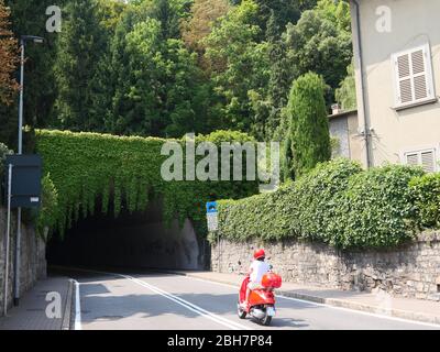 Bergamo, Italia - 06 agosto 2019: Una ragazza che guida uno scooter rosso Vespa nel bellissimo tunnel oscuro coperto di verde Foto Stock