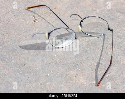 Un paio di occhiali con bordo in filo di titanio che si sono rotti e separati sulla sezione del ponte nasale mentre si posano su una superficie di cemento. Foto Stock