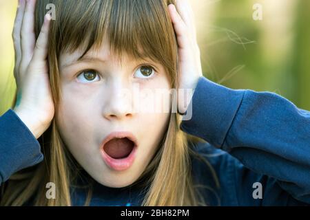 Ritratto di ragazza bambino sorpreso che tiene le mani alla sua testa all'aperto in estate. Scioccata femmina capretto in una calda giornata fuori. Foto Stock