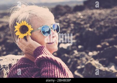 Bella donna anziana adulta con il fiore giallo del sole sulla testa prendere il sole e godere lo stile di vita in un luogo naturale all'aperto. Sorriso e felicità per donne mature con sfondo bokeh disfocused Foto Stock