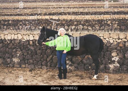 Bionda attraente europeo caucasico giovane donna di mezza età posa in piedi con un bel cavallo nero scuro. Sfondo da parete di campagna e concetto di stile di moda Foto Stock