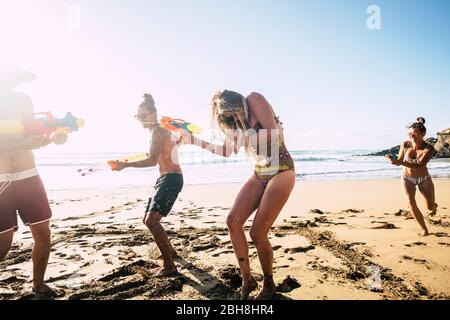 Persone che si divertono insieme in amicizia in spiaggia giocando con pistole ad acqua in bikini sotto il caldo sole estivo - pazzia e amici - immagine luminosa bella uomini e donne con mare in background Foto Stock