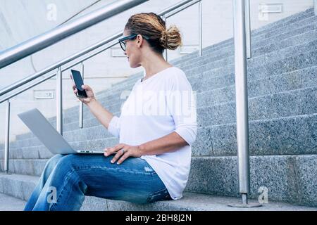 Donna d'affari bella donna che lavora con il telefono e computer portatile aoutdoor witting su una scala urbana in città - digitale nomade e freelance - libertà di lavorare all'aperto e godere di uno stile di vita moderno felice Foto Stock