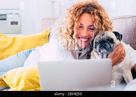 Felice bella giovane donna sorridere e guardare il computer portatile mentre abbracciare il suo vecchio divertente pug cane a casa sul divano - insieme per sempre e per concetto di terapia Foto Stock