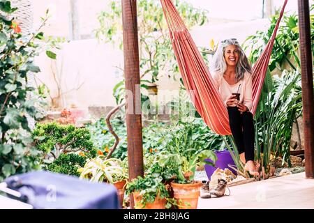 Allegro sorridente diversità concetto di gente con bella donna di tendenza adulto con capelli lunghi bianchi ridere sedersi su un'amaca a casa nel giardino bere un tè Foto Stock