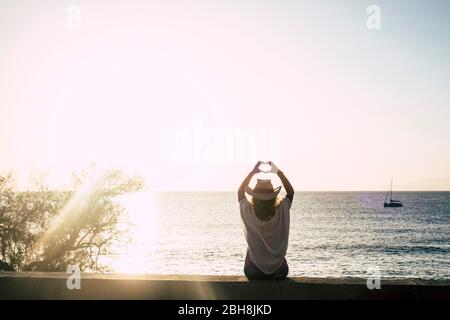 ragazza carina con cappello di stile estivo vigliottamente visto dalla parte posteriore facendo il segno del cuore con le mani - sole retroilluminazione e oceano con barca in background - libertà e godere di stile di vita concetto - cielo chiaro Foto Stock