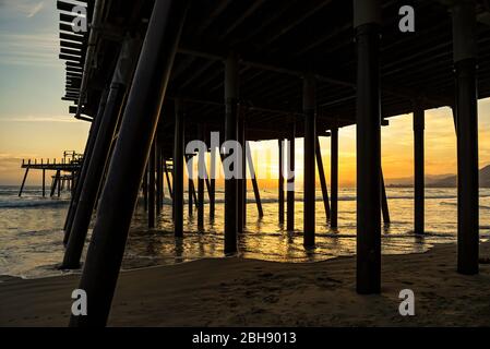 Blick durch den Pier in Pismo Beach von unten zum Pazifischen Ozean mit Surfern an der kalifornischen Westküste bei Sonnenuntergang Foto Stock